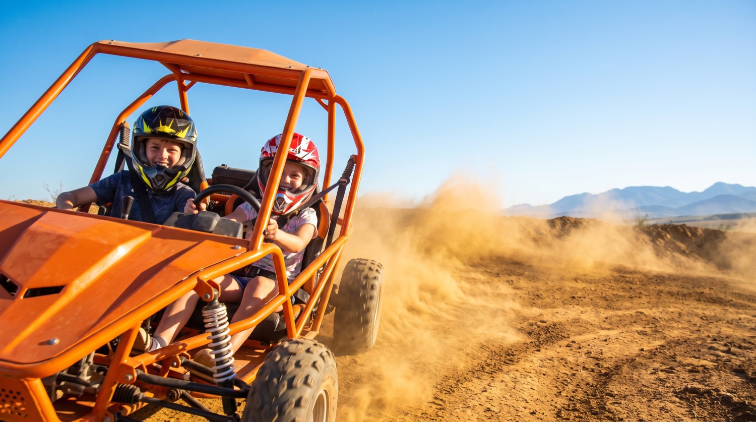 Two kids laughing in an orange two-seater buggy kicking up dust